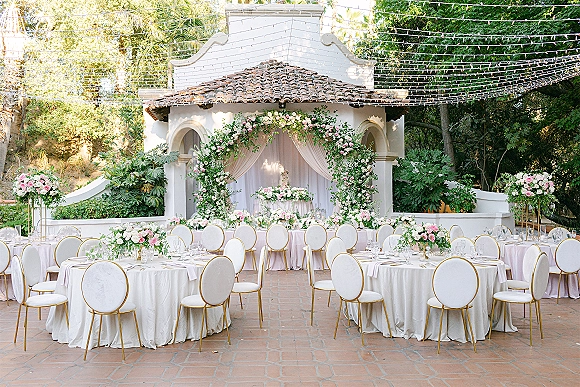 Reception tablescape with round white-linen tables, gold-rimmed chairs, floral centerpieces and string lights on a brick patio courtyard