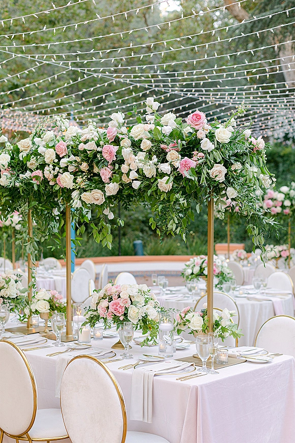 Reception tablescape with rose centerpiece and greenery garland under gold canopy and string lights, set on a garden patio with trees