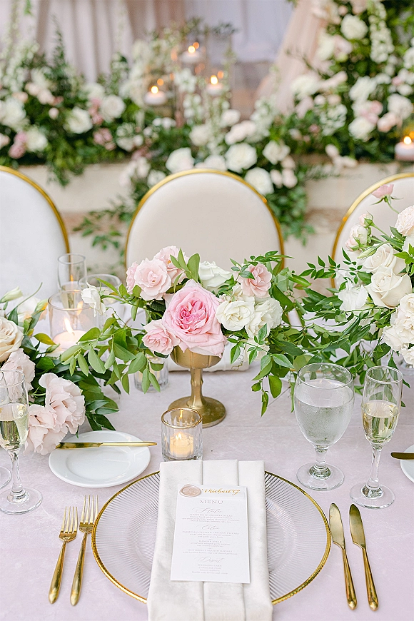 Reception tablescape with wedding head table decor, pink and white roses and greenery garland, gold flatware, candles, and white drapery backdrop