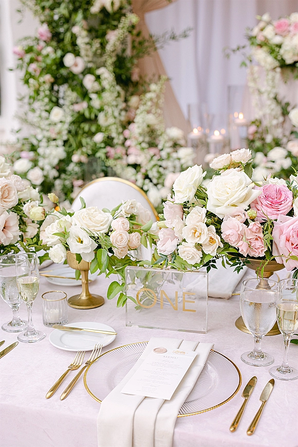 Reception tablescape with wedding table setting, pink and white rose centerpieces, gold flatware and candles on blush linen before drapery