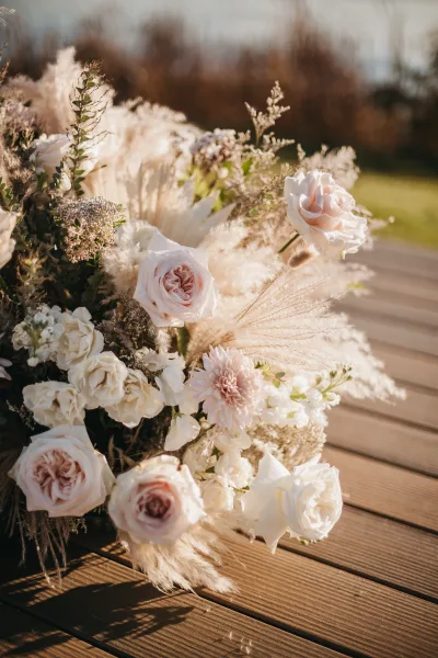 Wedding bouquet of blush rose bridal bouquet blooms with pampas grass accent, greenery, and dried grasses on a sunlit wood deck near trees