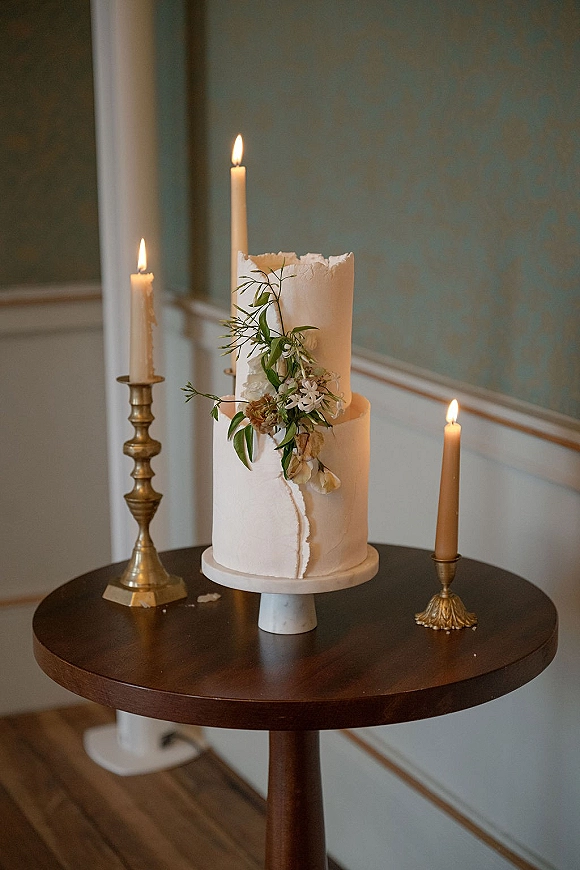 Wedding cake with textured buttercream and a floral topper on a pedestal table, flanked by brass taper candles against a paneled wall