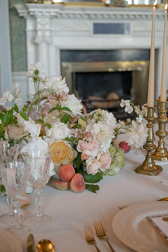 Reception tablescape with wedding table centerpiece of pastel roses and peonies, brass taper candles, crystal glassware, set by a fireplace mantel