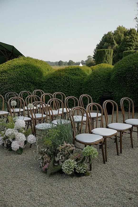 Outdoor ceremony seating with wood bistro chairs and white cushions lining a gravel courtyard aisle edged with low florals and boxwood hedges