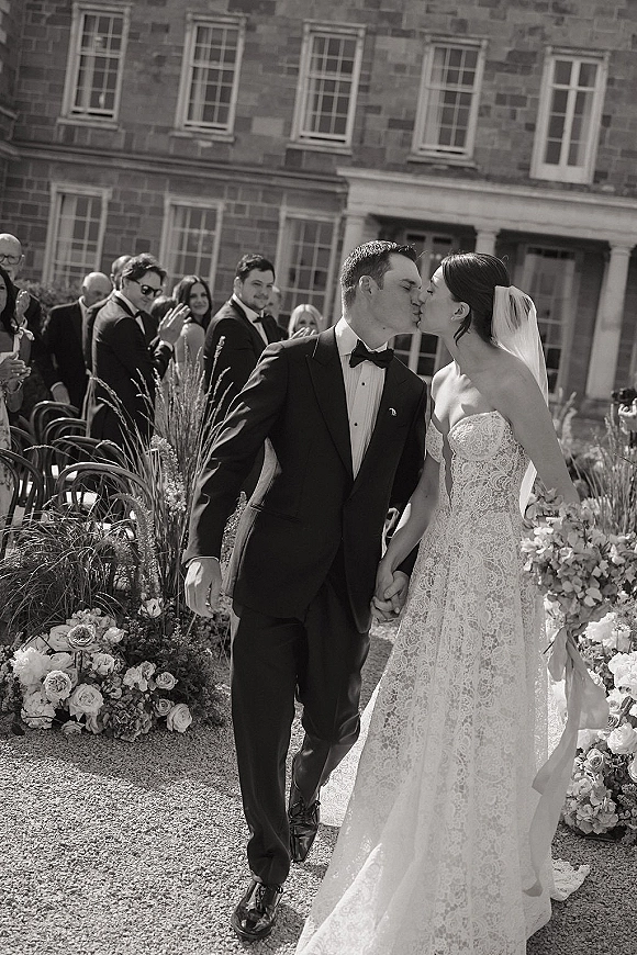 Wedding kiss portrait of bride and groom kiss holding hands in a courtyard aisle, her lace veil flowing past rose florals by stone facade