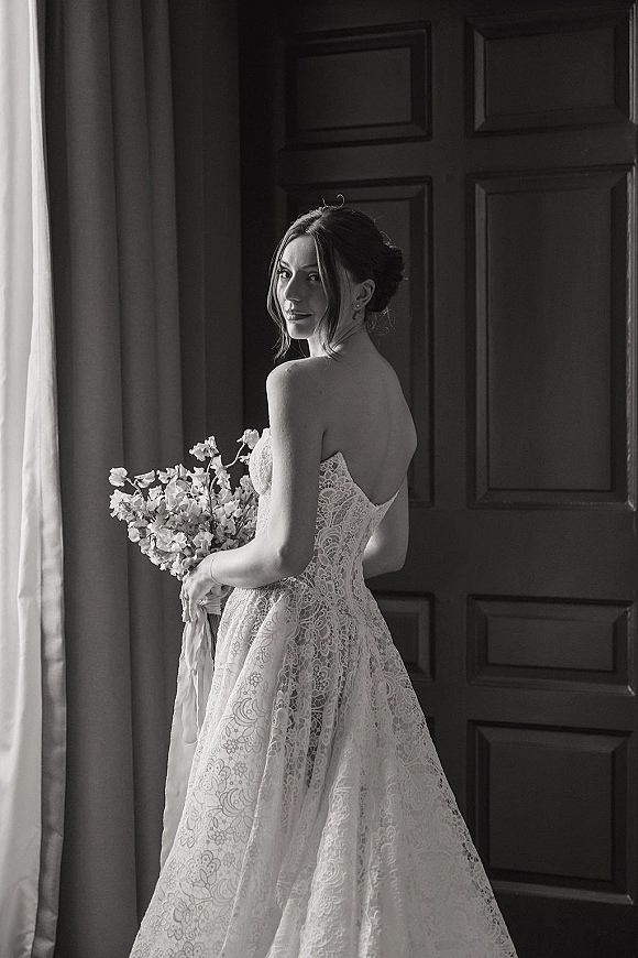 Bridal portrait in black and white of a bride holding bouquet, wearing a strapless lace wedding dress by a window with paneled doors