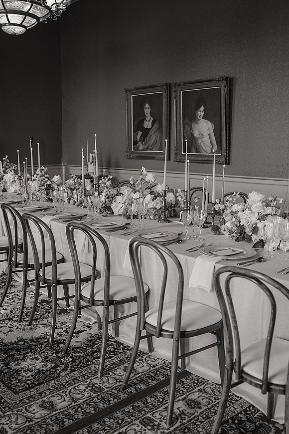 Reception tablescape with long banquet table decor, white linens, wedding table florals and taper candles under a chandelier amid vintage wallpaper