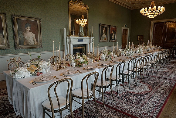 Reception tablescape with long banquet table decor on white linen, white floral centerpieces and brass candlesticks beneath chandeliers by a fireplace mirror
