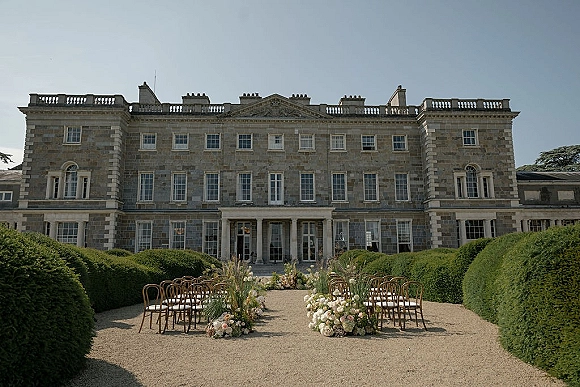 Outdoor ceremony setup with garden ceremony aisle lined by white rose and pampas florals, wood chairs in a gravel courtyard by a stone manor house