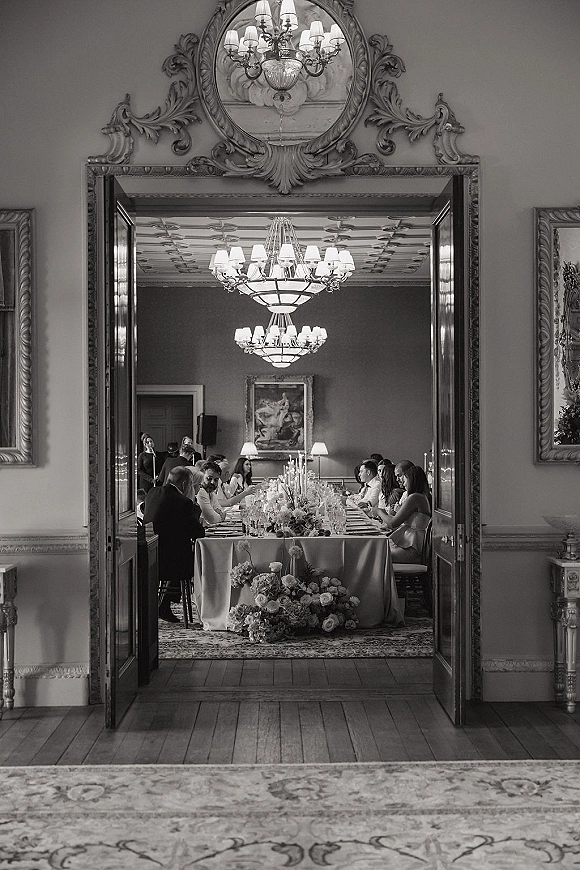Reception tablescape with a long banquet table, white tablecloth, taper candles, and low floral centerpiece beneath chandeliers in an ornate room