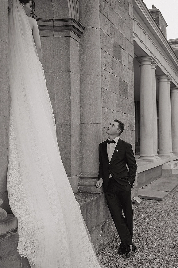 Couple portrait in a black and white wedding portrait, bride in lace gown and veil on stone steps as groom in tux looks up at her