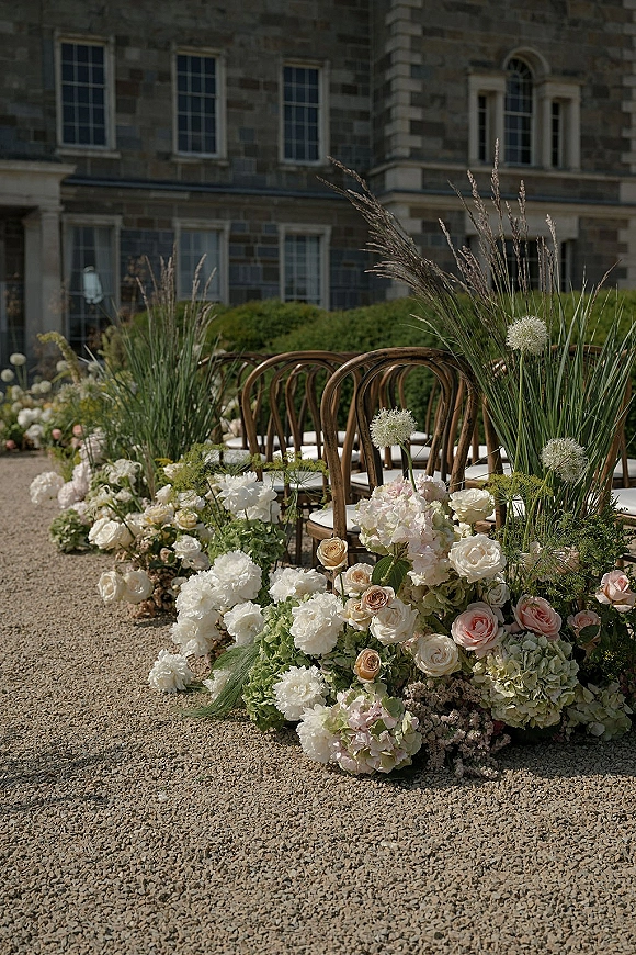 Ceremony aisle decor with a ground floral aisle of roses and hydrangea lining a gravel path beside wooden chairs at a stone estate facade