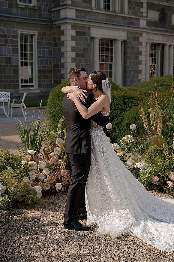 Wedding kiss as bride and groom embrace, her lace gown and veil trailing beside rose florals in a stone courtyard walkway