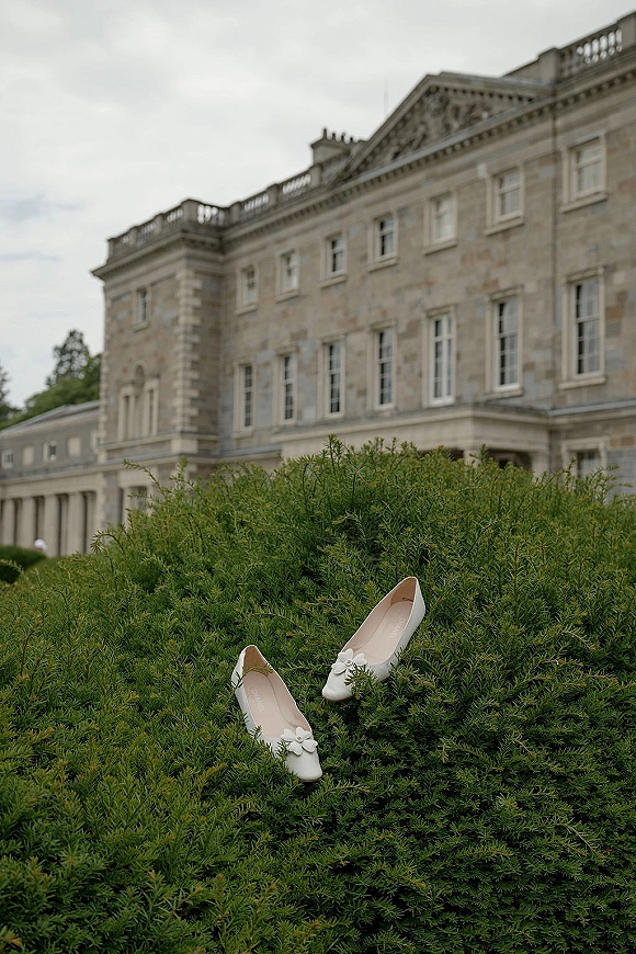 Wedding shoes, white bridal flats with floral appliqués resting on green shrubbery with a stone manor and cloudy sky behind