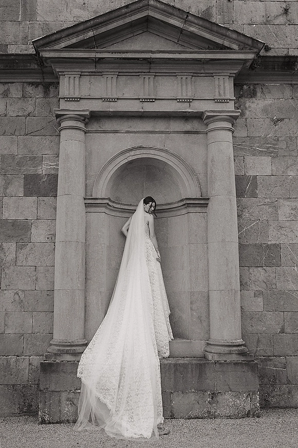 Bridal portrait in black and white of a bride looking over her shoulder, lace gown and cathedral veil trailing by stone columns and steps