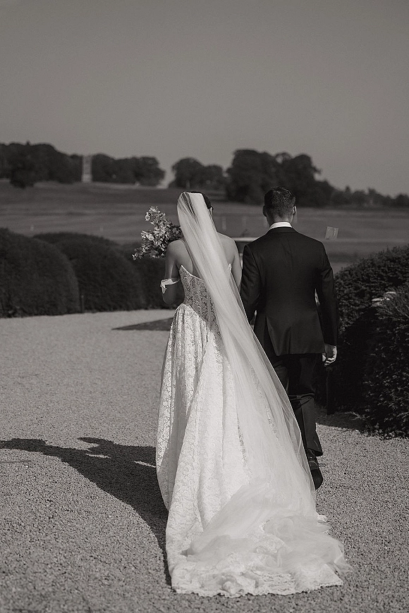 Wedding couple portrait of bride and groom walking away, her long veil and lace train flowing on a gravel path by hedges and field