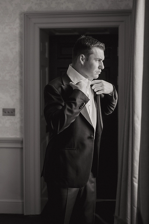 Groom portrait in window light as he adjusts his bow tie, wearing a tuxedo jacket with cufflinks, framed by a doorway and curtains