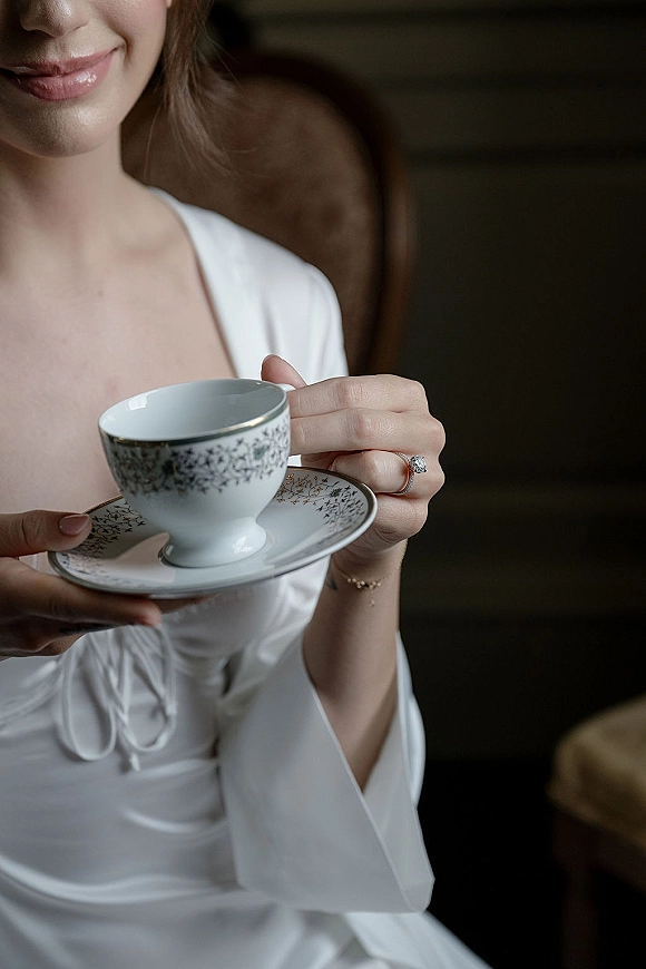 Bridal portrait of a bride holding teacup, showing her engagement ring and bracelet in a dark interior on an upholstered chair