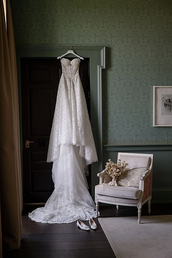 Wedding dress in lace with a sweetheart neckline hanging on a paneled door, with bridal shoes and bouquet on a chair nearby