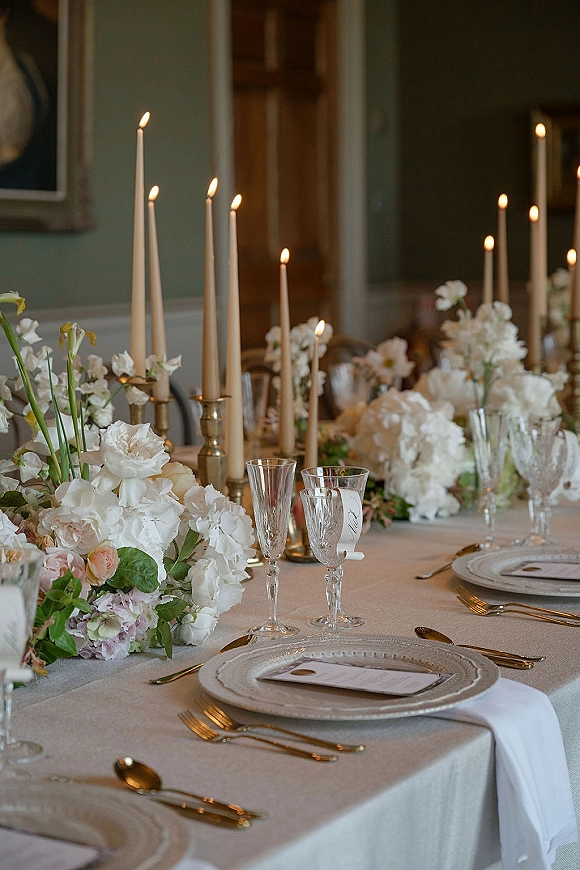 Reception tablescape with wedding table decor featuring white floral centerpiece, taper candles in brass holders, gold flatware, champagne flutes, green wall backdrop