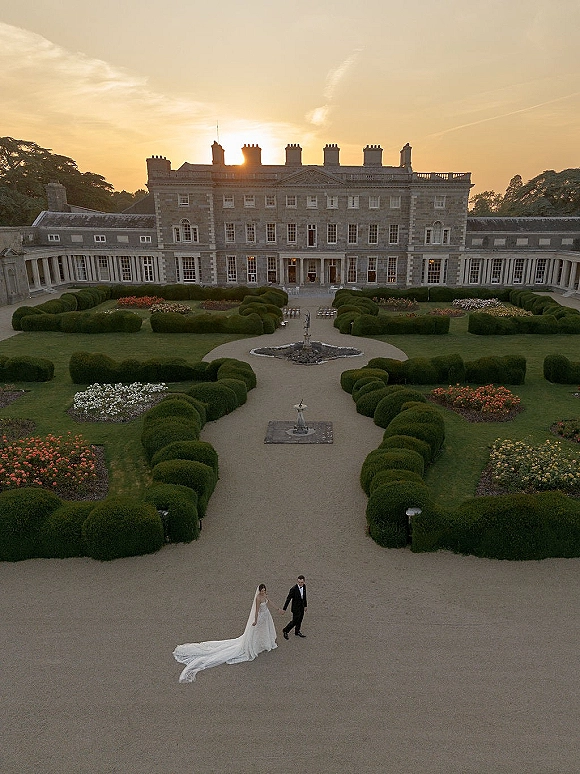 Couple portrait of bride and groom walking hand in hand, her veil and long train trailing on a gravel path in a manor garden at sunset
