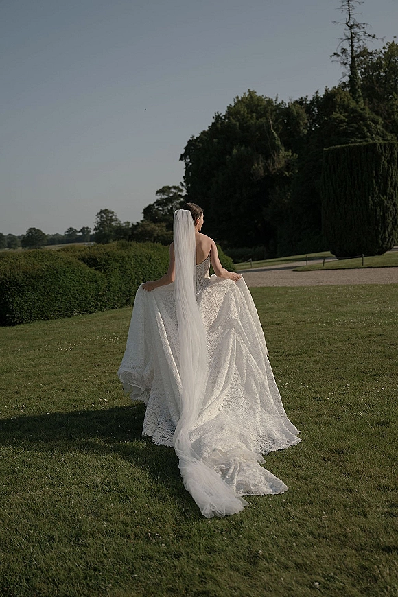Bridal portrait of a bride from behind in a lace strapless gown, holding her skirt as a long veil and train trail on a garden path