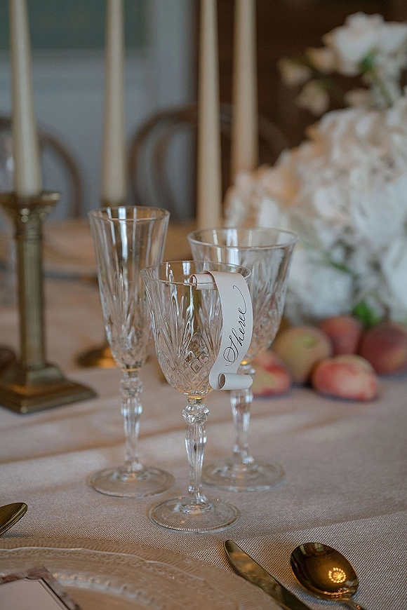 Wedding place card with calligraphy place card on a crystal champagne coupe, beside taper candles, white florals, peaches, and gold flatware on linen tablecloth