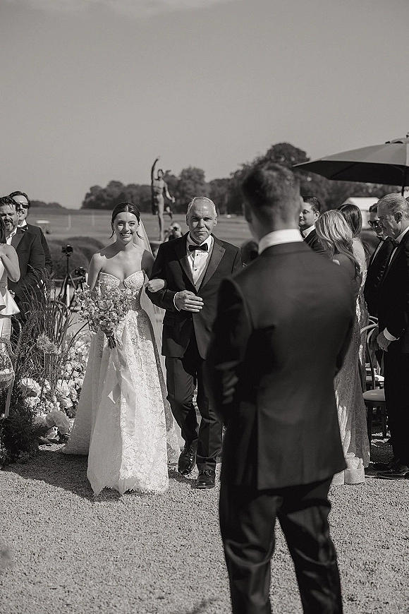 Wedding processional with bride walking down aisle beside her father, holding bouquet and veil, past seated guests on a tree-lined lawn