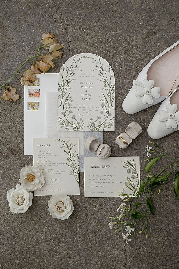 Invitation suite flat lay with arched wedding invitation, RSVP and envelopes, vintage postage, rings, and white floral heels on stone surface