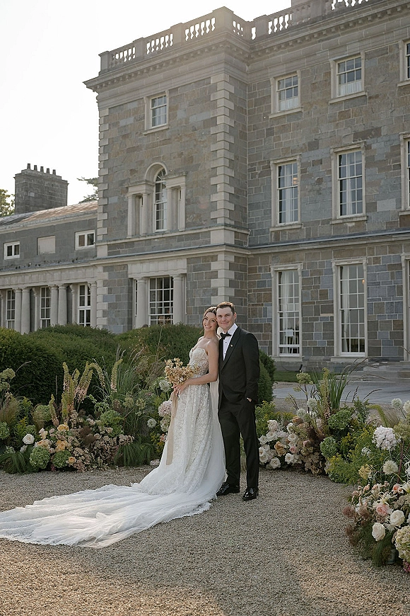 Couple portrait of bride in strapless lace gown with long train and veil beside groom in tux, holding bouquet before stone mansion facade