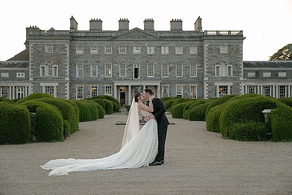 Wedding kiss portrait of bride and groom kissing, her veil and long train flowing beside his tuxedo on a gravel drive before a grand estate