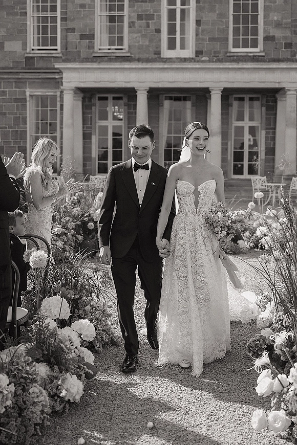 Wedding recessional as bride and groom walk the aisle holding hands, bride laughing in lace strapless gown and veil, manor garden path behind