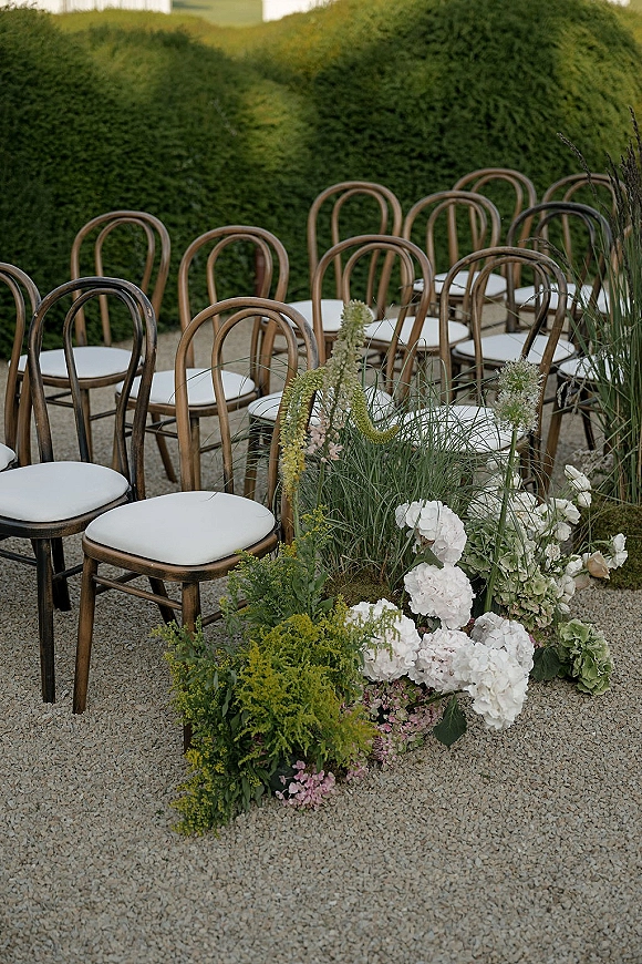 Ceremony seating with bentwood wedding chairs and white cushions beside a gravel aisle lined with low hydrangea flowers and hedges