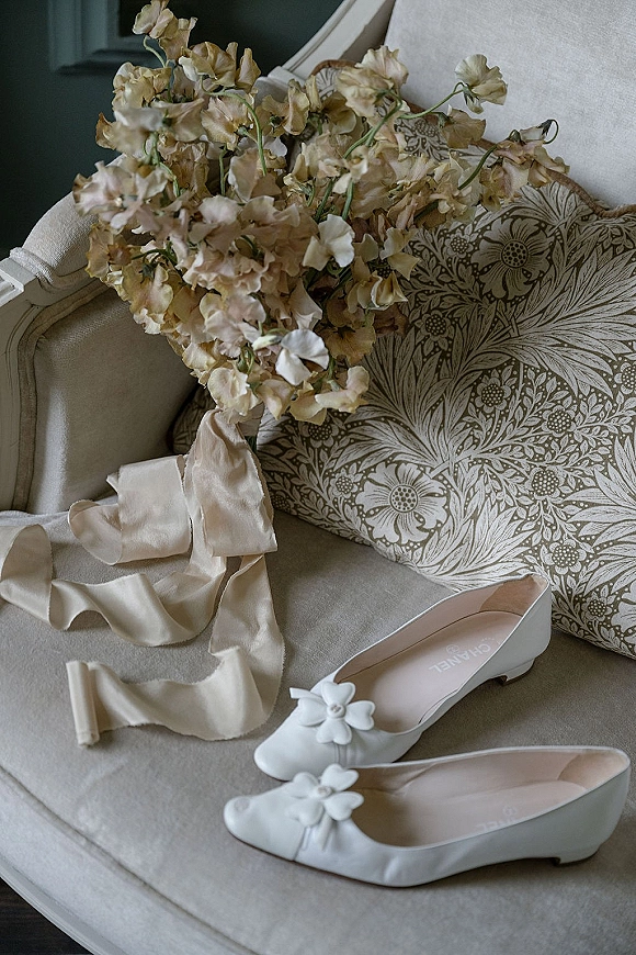 Bridal shoes, white bridal flats with flower appliques and ivory satin ribbon, styled with bouquet on an upholstered chair indoors