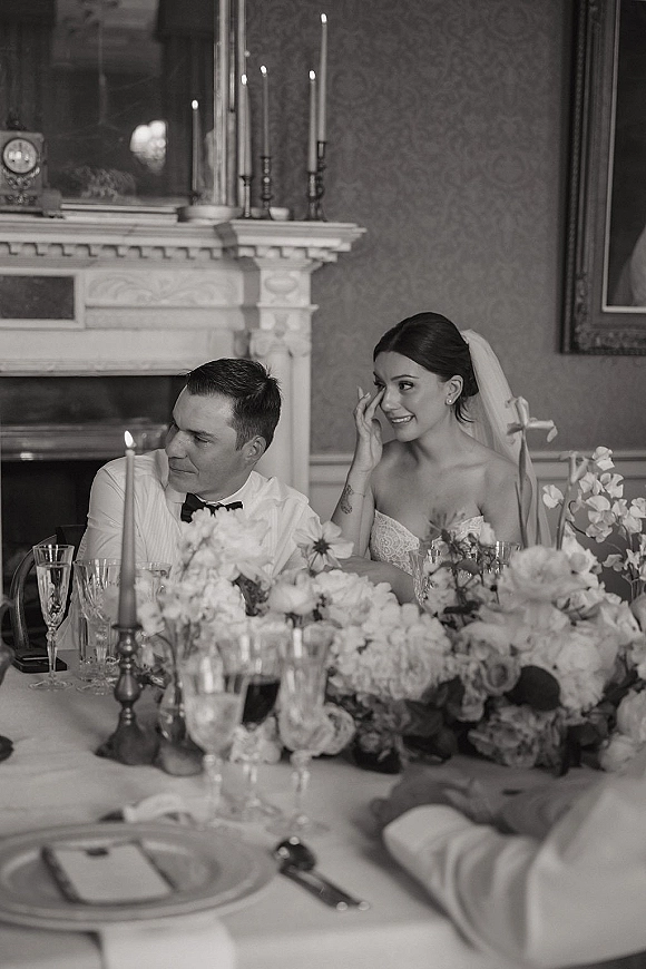 Wedding reception moment as bride wipes tears beside groom at candlelit sweetheart table with taper candles, floral centerpiece and fireplace backdrop