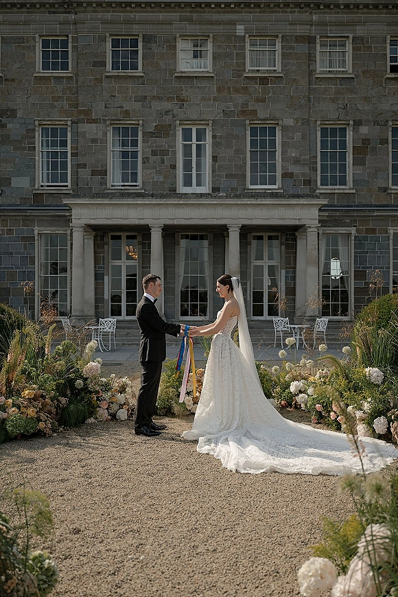 Ceremony moment as bride and groom hold hands beside ribbon wands, veil trailing in a gravel courtyard before a stone manor porch