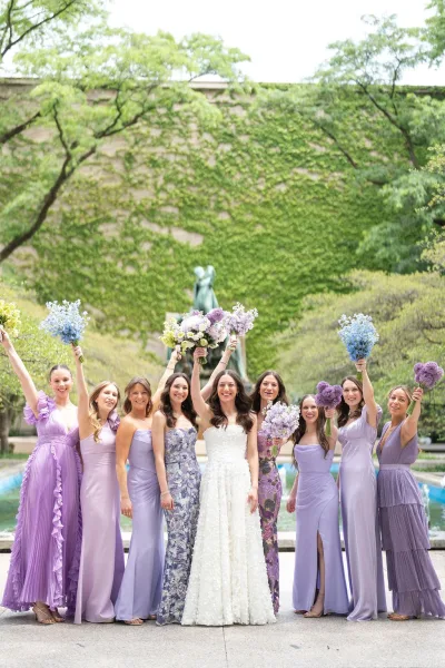 Bride and bridesmaids in mismatched bridesmaid dresses pose before an ivy wall, garden fountain and statue on stone pavement.