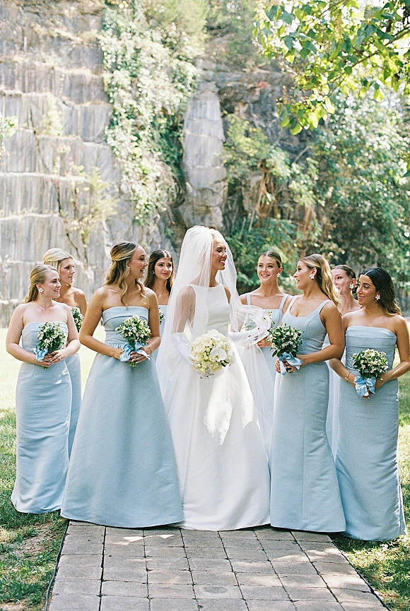 Bridal party portrait of bride with bridesmaids in light blue dresses holding green-and-white bouquets with blue ribbon wraps by a stone wall