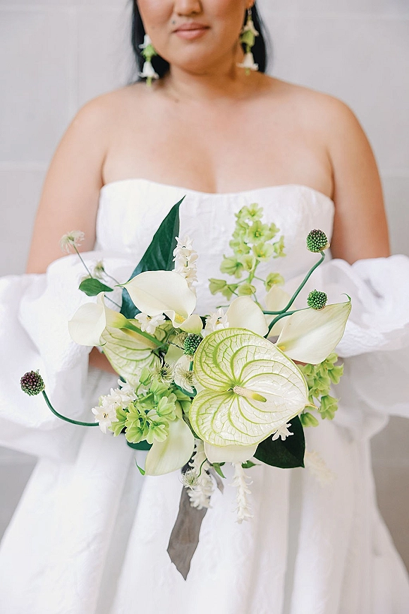 Bridal bouquet of white calla lilies and anthurium with lush greenery, held by a bride in a strapless gown against a clean white wall
