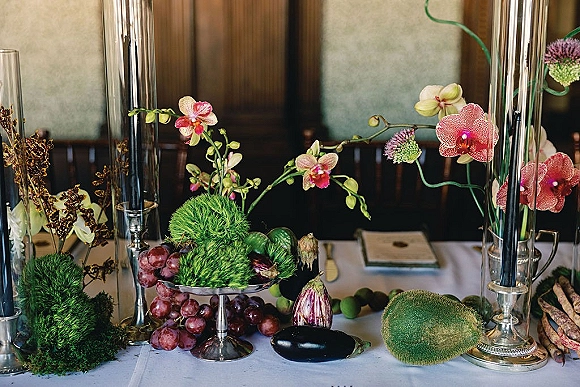 Reception tablescape with wedding table centerpiece of orchids, tall glass candle holders, and fruit and vegetables on a white tablecloth