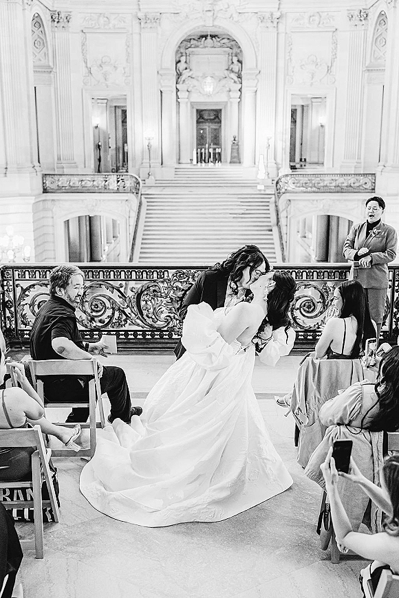 Ceremony kiss as two brides dip for a just married kiss, veil and long train flowing before a grand staircase and ornate balcony railing