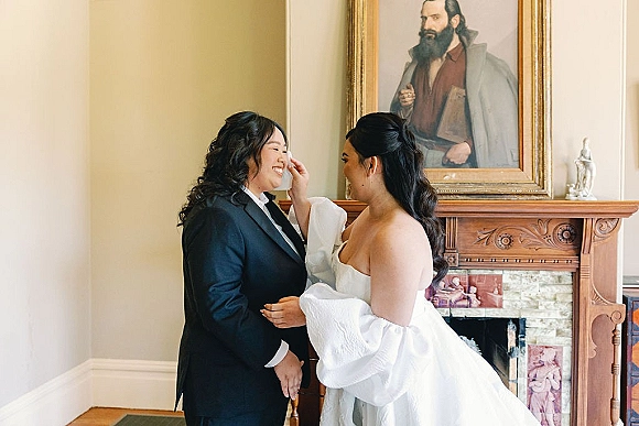Couple portrait of a same sex wedding couple, one bride in puff sleeve gloves adjusting her partner’s tie by a fireplace mantel