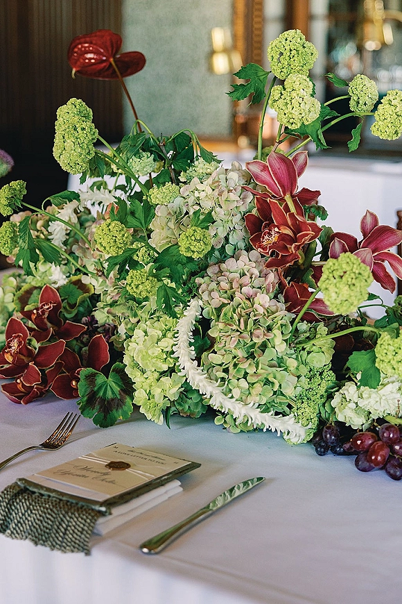 Wedding centerpiece with hydrangea centerpiece of green blooms, burgundy orchids and red anthurium on a white tablecloth with grapes and wax-seal place card
