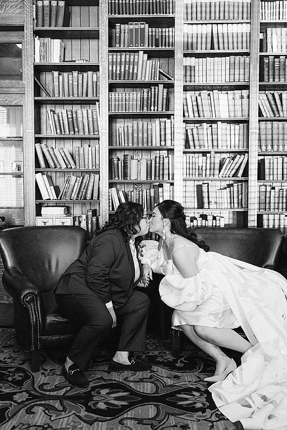Wedding kiss portrait of bride and groom kissing on a leather sofa, her veil draped, with library bookshelves and a patterned rug behind them