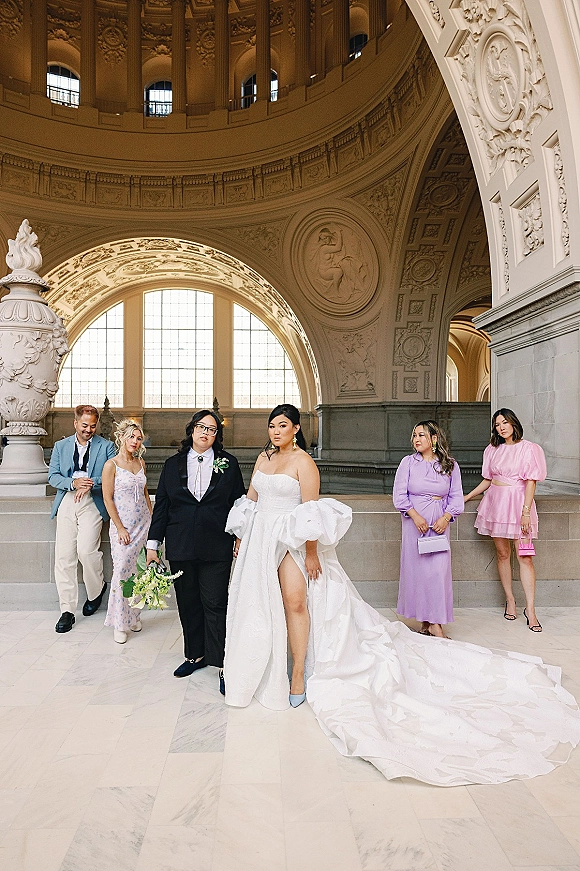 Wedding party portrait with bride in strapless gown and groom in black suit, bridesmaids with bouquets on marble steps in arched hall.