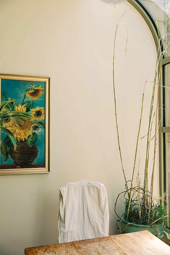 Wedding dress on chair beside a framed painting and potted plant, set by an arched window in a softly lit indoor room