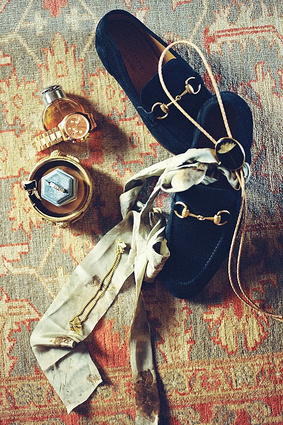 Groom accessories flatlay with velvet loafers, gold watch, cologne, ring box, pocket watch and silk tie on a patterned vintage rug