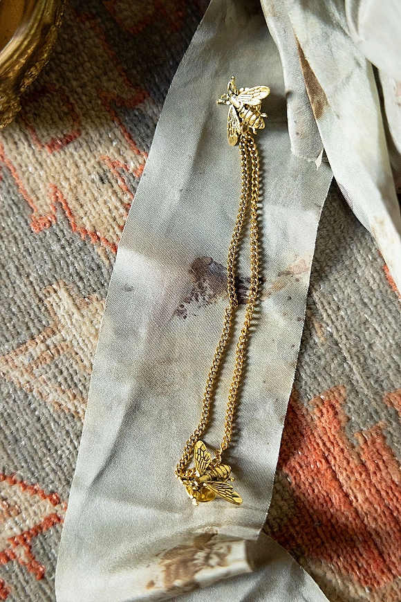 Bridal jewelry featuring a gold bee necklace with layered pendants and ring, styled on a white ribbon atop a patterned rug flatlay