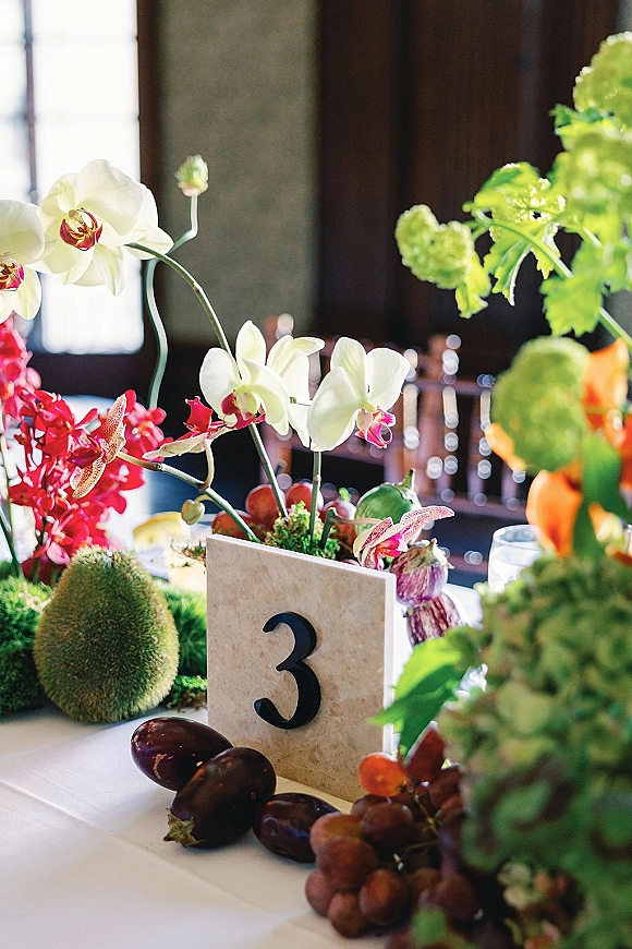 Reception tablescape with wedding table centerpiece of orchids, red and green florals, grapes and eggplant, plus table number card in window light