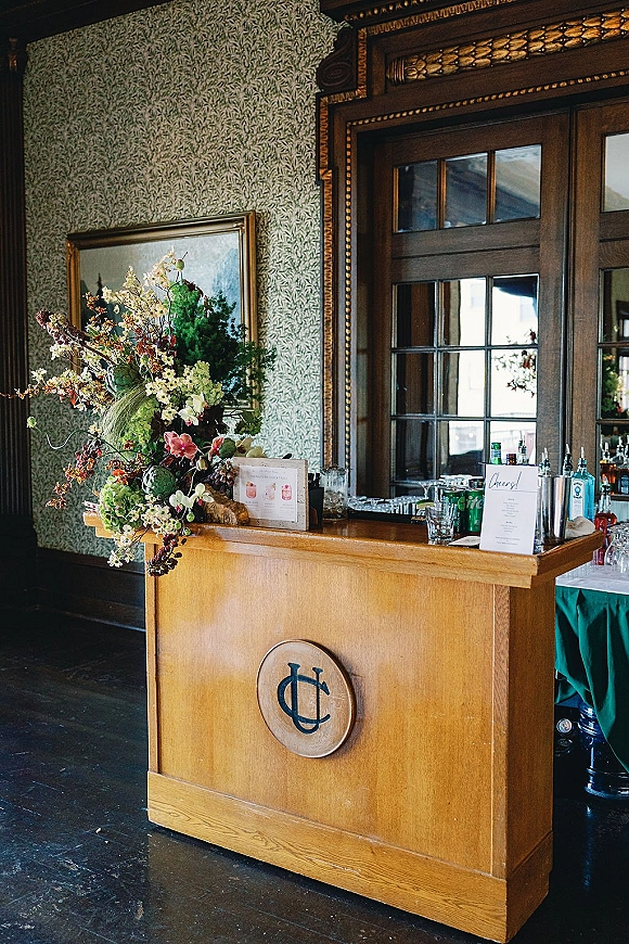 Wedding bar setup with a mobile wedding bar cart, cocktail menu sign, liquor bottles and glassware beside wood-paneled windows in a classic room